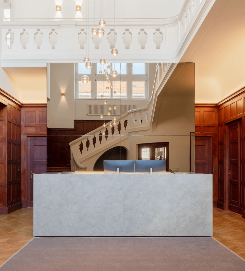 Ein hellgrauer Empfangstresen in einer prächtigen Halle mit dunklen Holzvertäfelungen, der eine Treppe und moderne Kugelleuchten spiegelt.