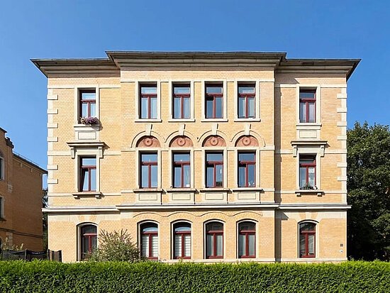 Symmetrisches gelbes Backsteingebäude mit dunkelroten Fenstern, Zierbögen und einer grünen Hecke unter blauem Himmel.