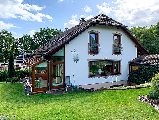 Ein charmantes weißes Haus mit Wintergarten, verzierten Fassadenmalereien und einem gepflegten grünen Garten unter bewölktem Himmel.
