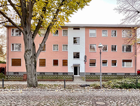 Lachsrosa Gebäude mit weißem Mittelteil, gerahmt von großem Baum mit gelben Herbstblättern und Laterne auf Kopfsteinpflaster.
