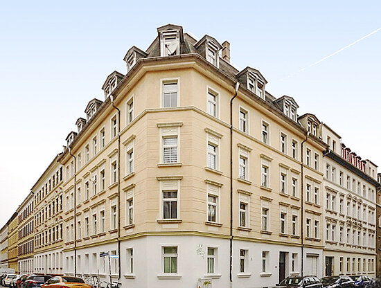 Ansicht eines großen, hellbeigen historischen Eckgebäudes mit vielen Fenstern und Dachgauben, an einer Stadtstraße mit Autos.