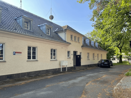 Cremefarbenes Gebäude mit Gauben, dunklem Dach, dunklem Sockel und einem schwarzen SUV davor geparkt.