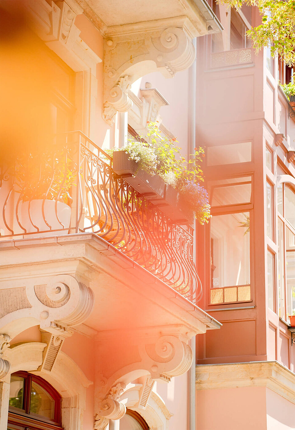 Rosafarbene Fassade mit verziertem Balkon, schmiedeeisernem Geländer und bunten Blumenkästen im Sonnenlicht.