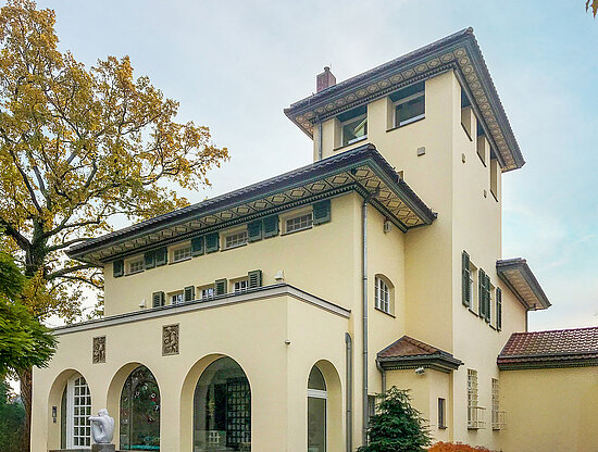 Große Jugendstilvilla mit Turm, cremefarbener Fassade, grünen Fensterläden und einem Baum mit goldenem Herbstlaub.