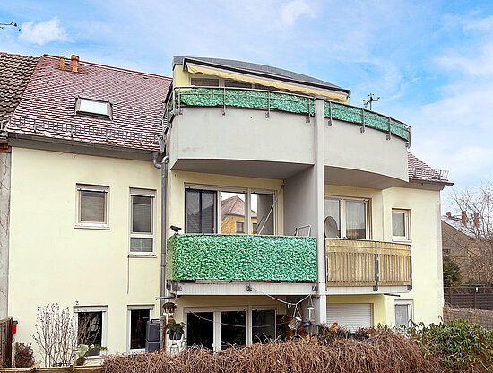 Fassade eines Wohngebäudes mit zwei Balkonen. Verfügt über grünen Sichtschutz, ein rotes Ziegeldach und einen Vogel auf dem Geländer.