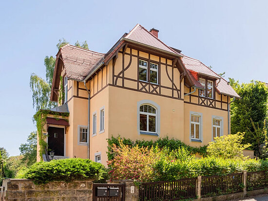 Charmantes gelbes Haus mit braunem Fachwerk, rotem Dach und grüner Bepflanzung unter blauem Himmel.