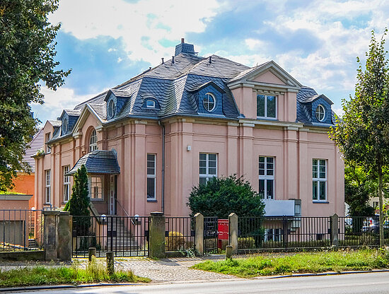 Elegante hellrosa Villa mit dunkelgrauem Mansardendach, Dachgauben und eingezäuntem Eingang, vor blauem Himmel.