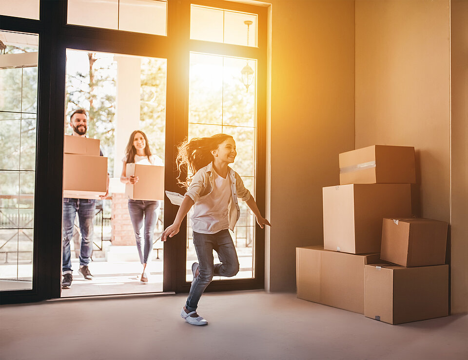 Familie zieht mit Umzugskartons in ein neues, helles Zuhause ein, während ein Kind fröhlich durch den leeren Raum läuft