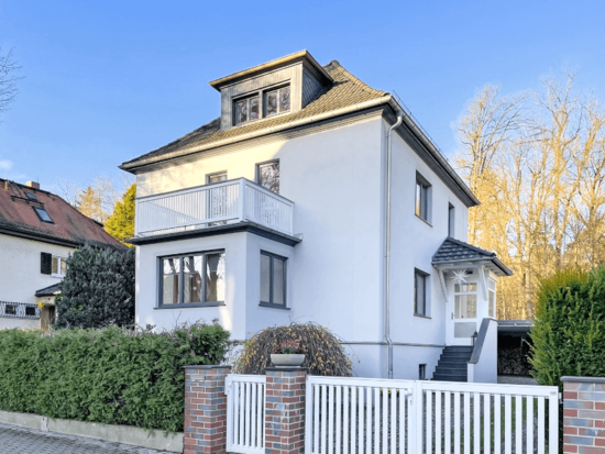 Weißes zweistöckiges Haus mit Balkon, grauem Dach, dunklen Fenstern, weißem Zaun, grüner Hecke und Bäumen unter blauem Himmel.