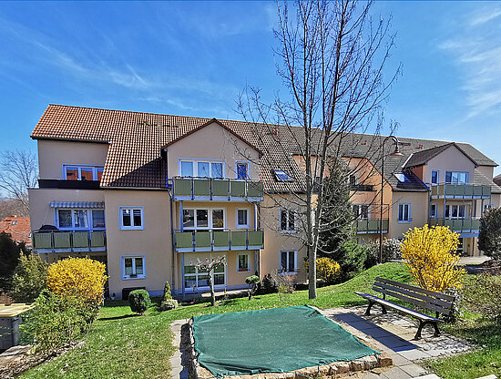 Wohngebäude mit gelben Fassaden, roten Dächern, grünen Balkonen und blühenden gelben Büschen unter blauem Himmel.