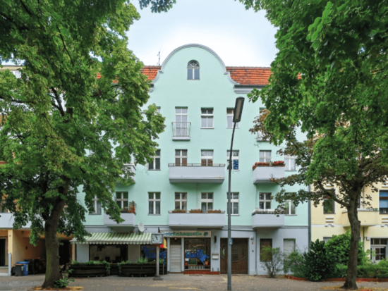 Ein minzgrünes Mehrfamilienhaus mit Balkonen und einem "Getränkequelle"-Laden, umrahmt von grünen Bäumen an einem sonnigen Tag.