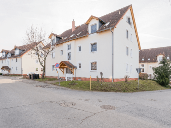 Weiße Wohnhäuser mit roten Dächern, Gauben und überdachten Eingängen an einer Straße mit kahlem Baum.