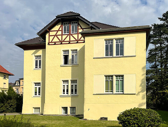 Helles gelbes Haus mit braunem Fachwerkgiebel, dunklem Dach, vielen Fenstern, grünem Rasen und hohem Baum.