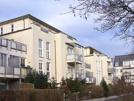 Reihe cremefarbener Mehrfamilienhäuser mit Balkonen, grünen Sträuchern und kahlen Bäumen unter bewölktem Himmel.