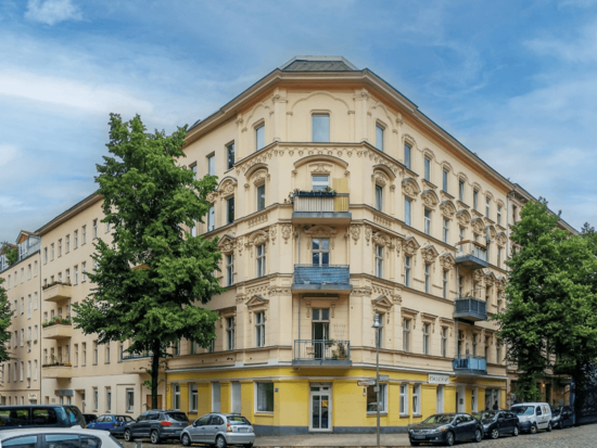 Ein hellgelbes, verziertes Eckgebäude mit blauen Balkonen und großem Baum an einer Stadtstraße mit parkenden Autos.