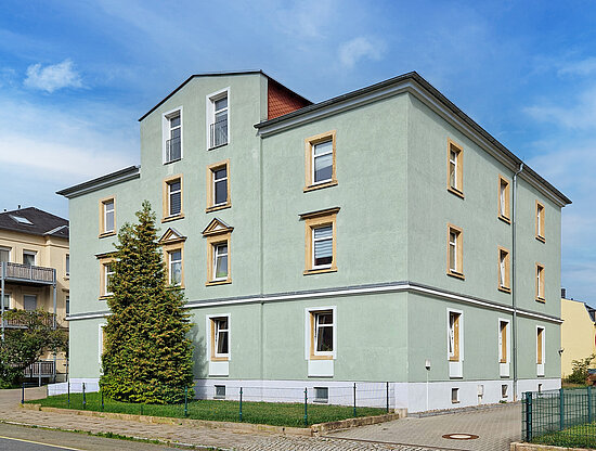Mehrstöckiges hellgrünes Gebäude mit vielen Fenstern, einem kegelförmigen Baum davor und blauem Himmel.