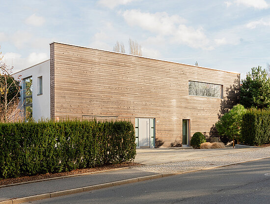 Modernes Haus mit horizontaler Holzverkleidung, weißen Wänden, großen Fenstern, grüner Hecke und gepflasterter Einfahrt.
