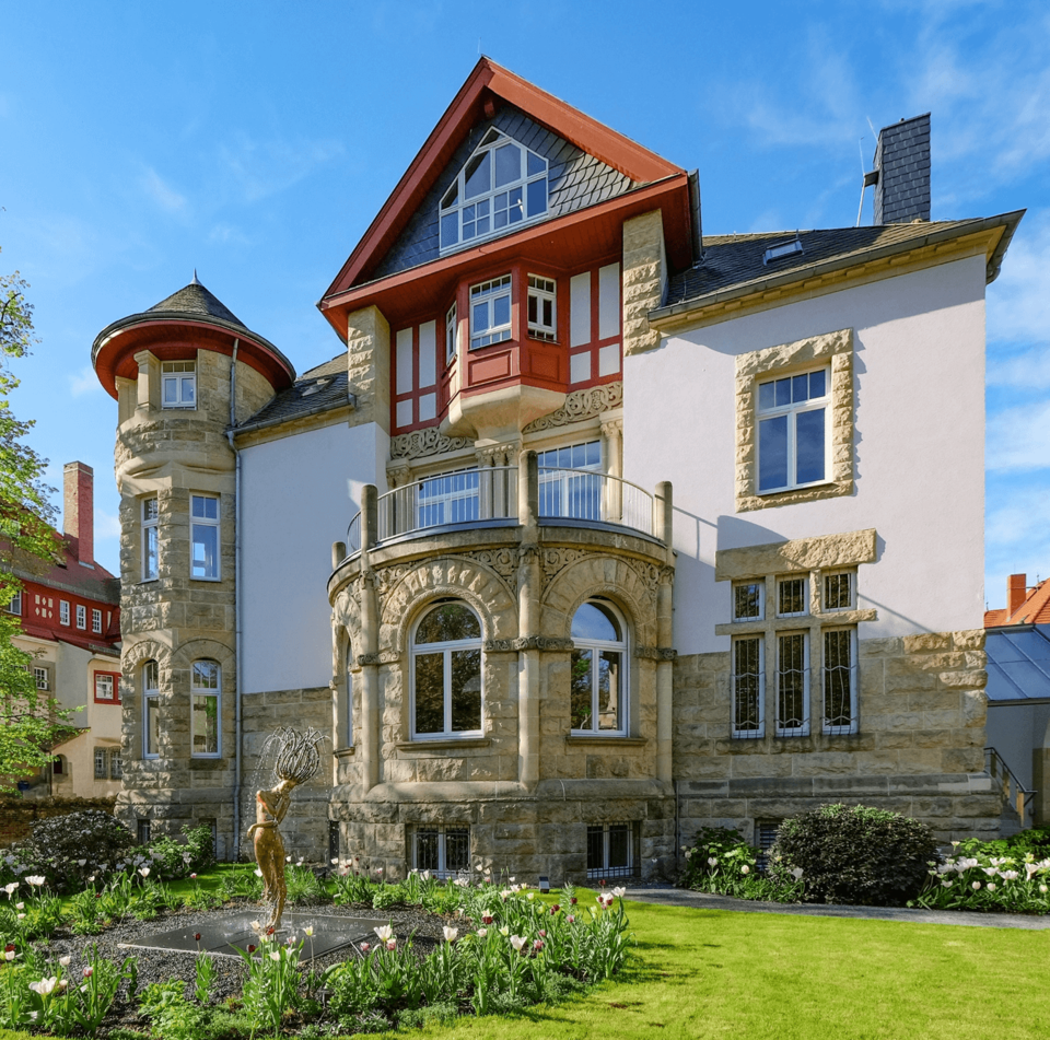 Verzierte historische Villa mit Steinfassade, Rundturm, roten Giebeln, Garten mit Tulpen und Brunnenskulptur.