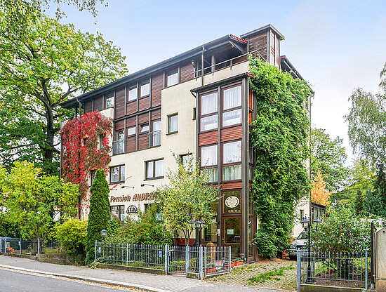 Außenansicht der Pension Andrea, ein Gebäude mit rotem und grünem Efeu, umgeben von Herbstbäumen und einem Zaun.