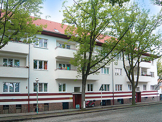 Weißes Mehrfamilienhaus mit rotem Dach, Balkonen und rot gestreiftem Sockel, umrahmt von grünen Bäumen an einer Kopfsteinpflasterstraße.