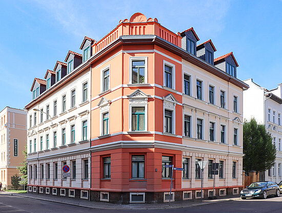 Eckgebäude mit orangefarbenen und cremefarbenen Fassaden, Gauben und Straßenschildern unter blauem Himmel.