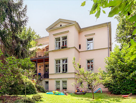 Außenansicht eines großen, hellrosa Hauses, umgeben von einem üppigen grünen Garten mit Bäumen und Kinderspielzeug.