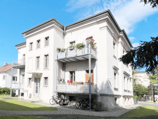 Helles klassisches Gebäude mit Balkonen und roten Blumen, Fahrrädern davor, grünem Rasen und blauem Himmel.