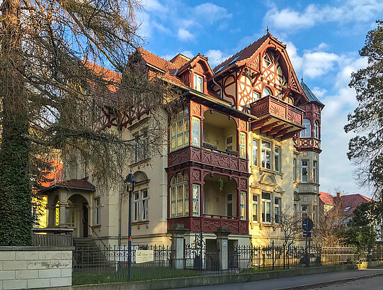 Historische Villa mit beiger Fassade, rotbraunen Balkonen, Fachwerkgiebeln und Turm unter blauem Himmel.