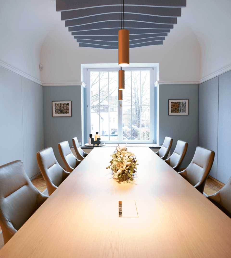 Moderner Konferenzraum mit langem Holztisch, beigen Stühlen, orangefarbenen Pendelleuchten und wellenförmiger Decke.