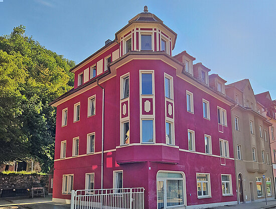 Ein helles Magenta-Gebäude mit achteckigem Turm und weißer Verkleidung, neben älteren Gebäuden und einem grünen Hügel.