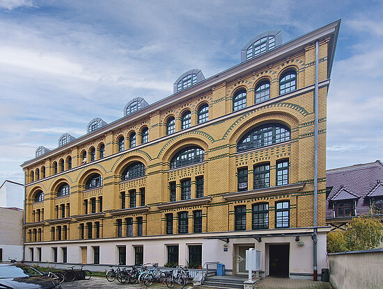 Gelbes Backsteingebäude mit keilförmigem Ende, Bogenfenstern und grünen Akzenten unter bewölktem Himmel. Fahrräder geparkt.