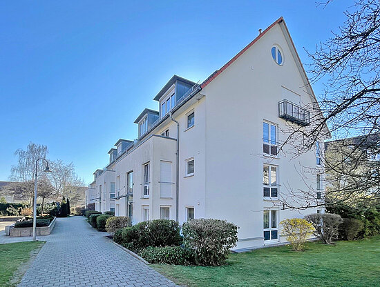 Ansicht moderner Mehrfamilienhäuser mit weißen Fassaden, Giebeldächern und Gauben unter blauem Himmel, neben einem gepflasterten Weg und Grünflächen.