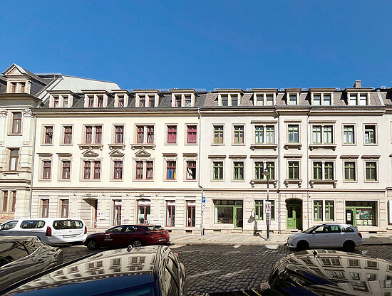 Straßenansicht historischer Mehrfamilienhäuser mit farbigen Fenstern, Autos auf Kopfsteinpflaster unter blauem Himmel.