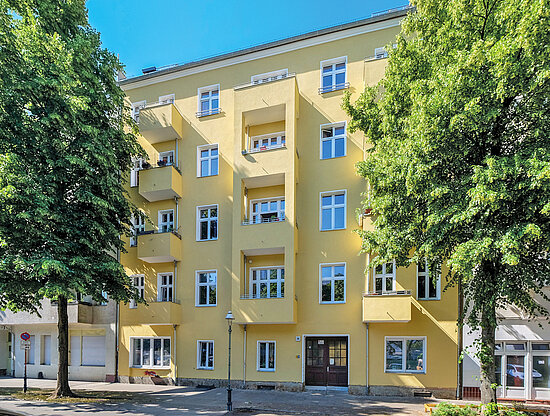 Außenansicht eines hellgelben Mehrfamilienhauses mit vielen Fenstern und Balkonen, umrahmt von zwei großen grünen Bäumen.