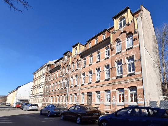 Blick auf eine Straße mit traditionellen Backsteinwohnhäusern unter blauem Himmel, Autos parken am Straßenrand.