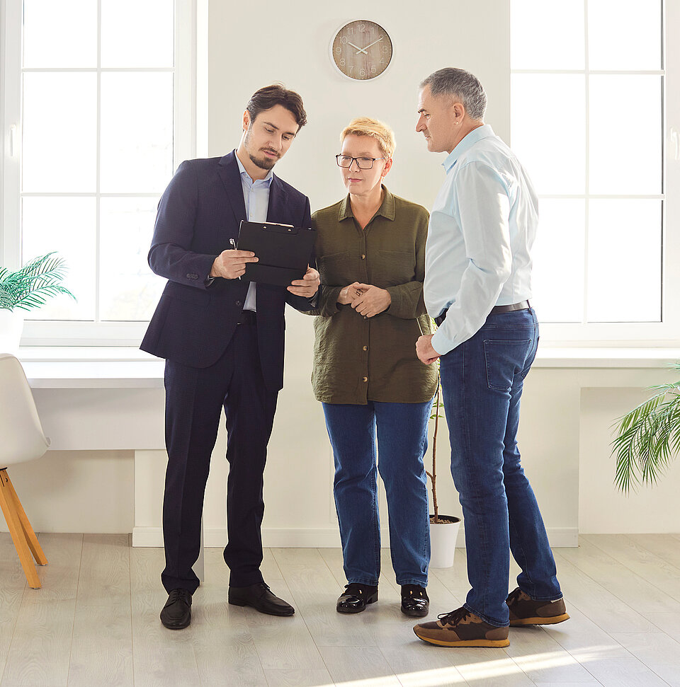 Ein Makler im Anzug erklärt einem Ehepaar Dokumente auf einem Klemmbrett in einem hellen Raum mit Fenstern.