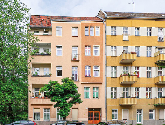 Zwei farbige Mehrfamilienhäuser, eines beige mit rotem Dach, das andere gelb-weiß gestreift mit Balkonen, grüne Bäume.