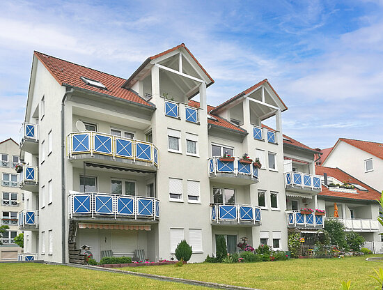 Modernes Apartmentgebäude mit hellgrauen Wänden, orangefarbenen Ziegeldächern und blauen Balkonen mit weißem X-Muster.