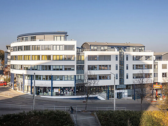 Modernes weißes Gebäude mit geschwungener Fassade, Glasaufzug und Erdgeschossläden, inkl. Norma Supermarkt.