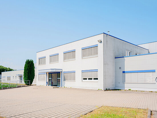 Modernes weißes Gebäude mit blauer Zierleiste, Außenjalousien, gepflastertem Hof und hohem Baum unter blauem Himmel.