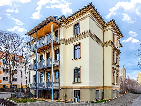 Ein cremefarbenes, klassisches Gebäude mit modernem dreistöckigem Balkonanbau unter teilweise bewölktem Himmel.