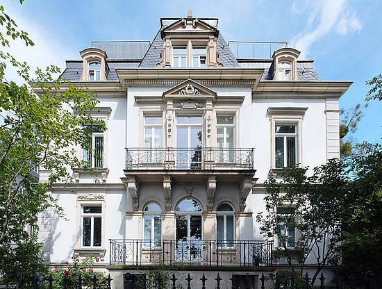Frontfassade einer eleganten weißen historischen Villa mit Balkonen, Zierelementen, Mansarddach und grünen Bäumen.