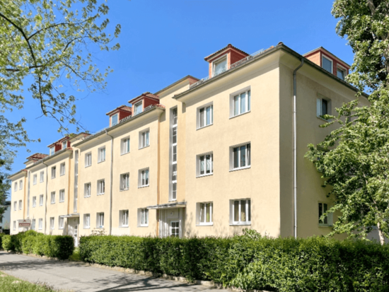 Langes, cremefarbenes Wohnhaus mit Gaubenfenstern und grüner Hecke unter klarem blauem Himmel.