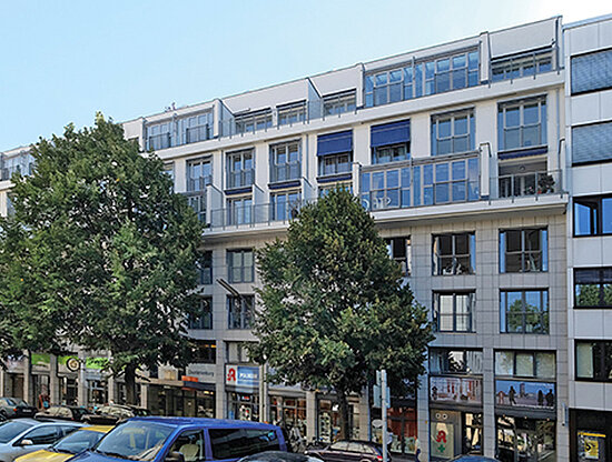 Außenansicht eines modernen Mehrfamilienhauses mit Balkonen, Geschäften im Erdgeschoss und grünen Bäumen.