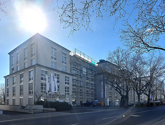 Großes Gewerbegebäude mit "MULTIPOLSTER"-Beschriftung, teilweise in Renovierung, an hellem Tag mit kahlen Bäumen.