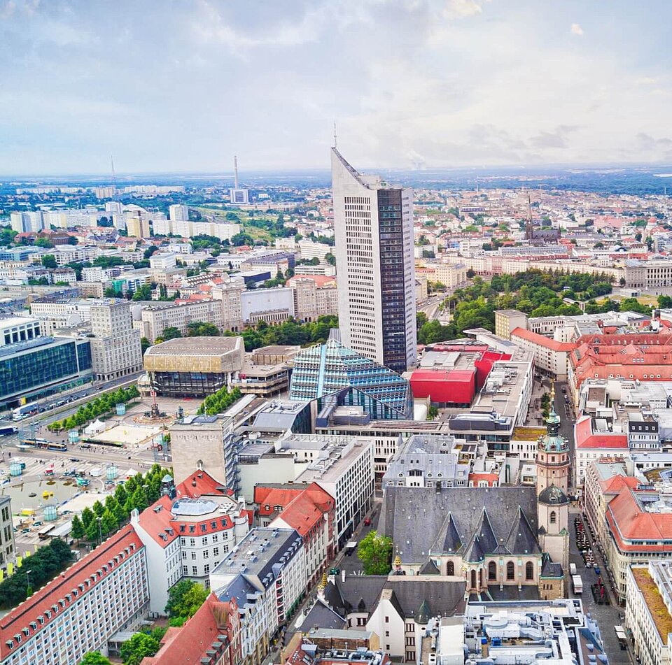 Luftaufnahme von Leipzig mit City-Hochhaus, gläsernem Paulinum und Thomaskirche unter bewölktem Himmel.