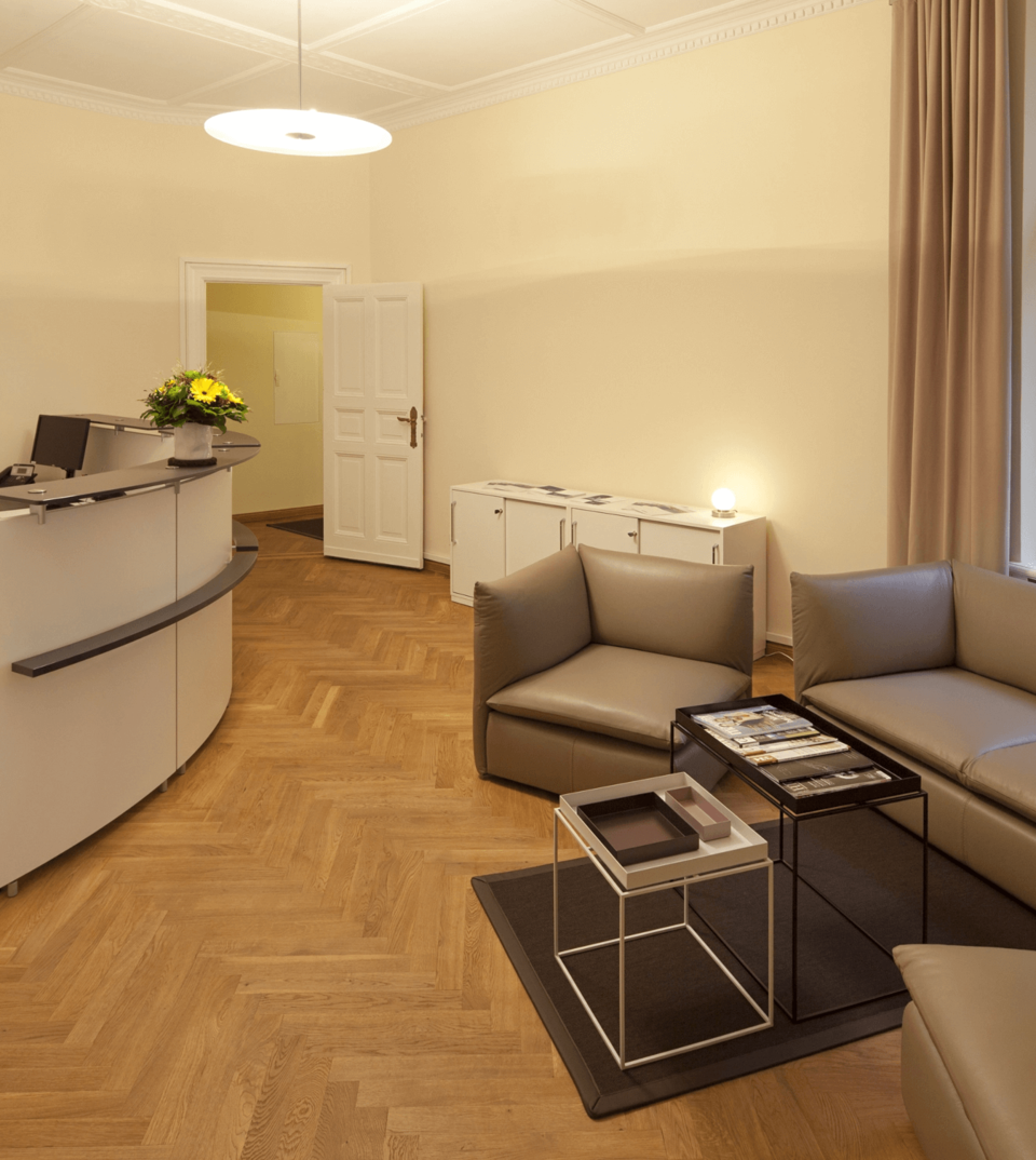 Heller, moderner Bürobereich mit weißem Empfangstresen, beigen Ledersitzen, Fischgrätparkett und Pendelleuchte.