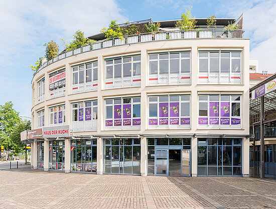 Vorderansicht eines cremefarbenen Geschäftsgebäudes mit Geschäften, "Zu vermieten"-Schildern und Dachgarten unter blauem Himmel.