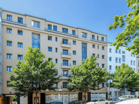 Außenansicht eines modernen, hellbeigen Mehrfamilienhauses mit grünen Bäumen, Balkonen und Geschäften im Erdgeschoss an einem sonnigen Tag.