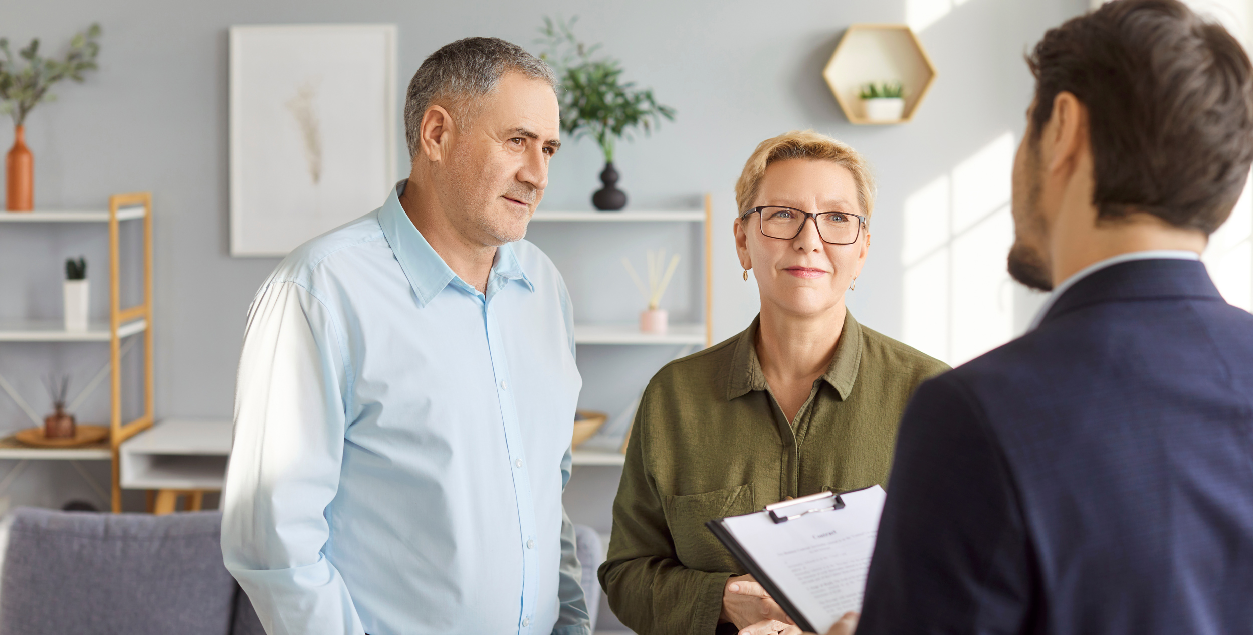 Ein Eigentümer-Paar hört einem männlichen Makler mit einem Klemmbrett in der Hand aufmerksam zu. Sie befinden sich in einem modernen Raum.